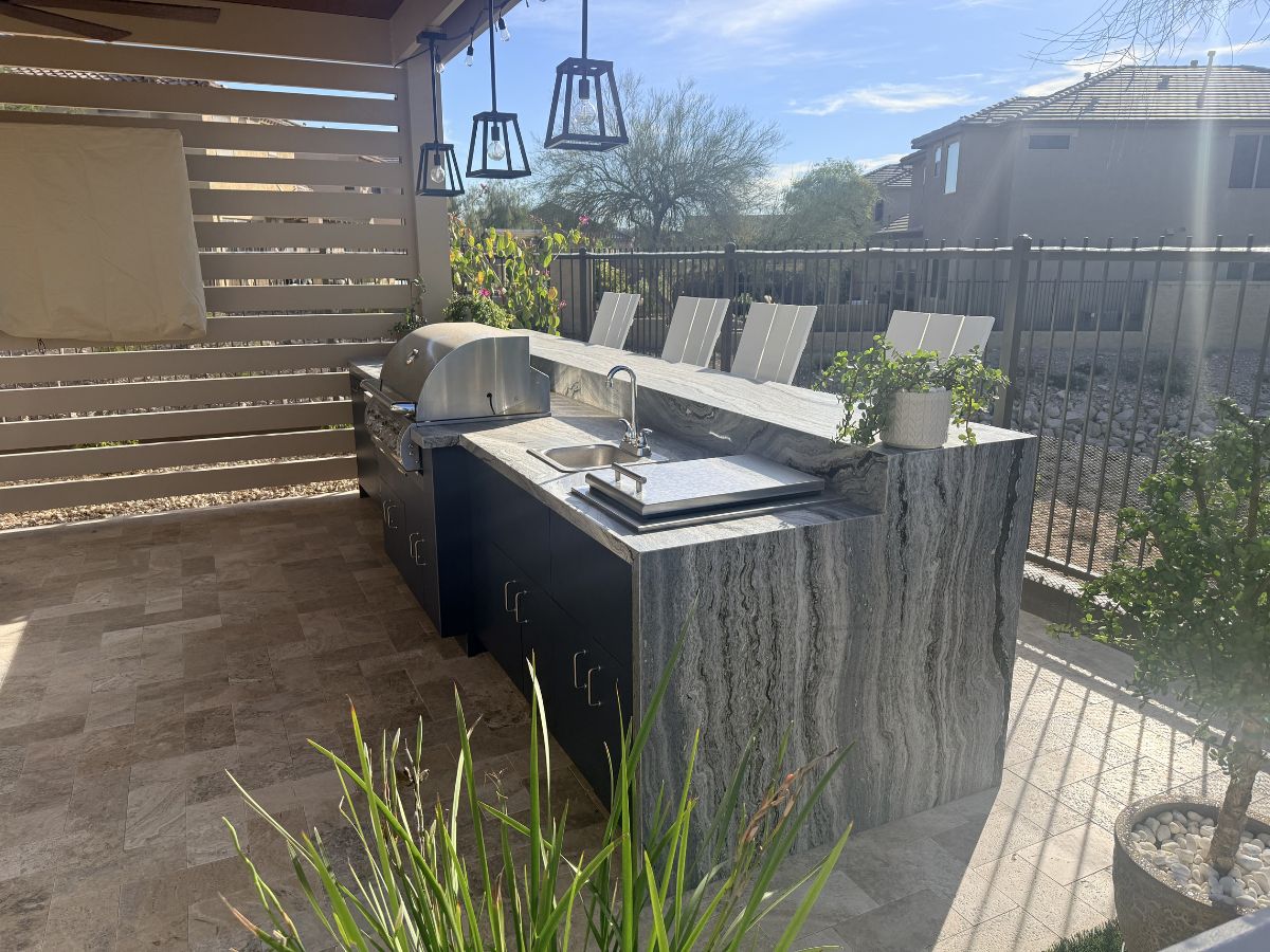 Modern outdoor kitchen with waterfall stone island, built-in stainless grill, sink, and bar seating on sunny patio