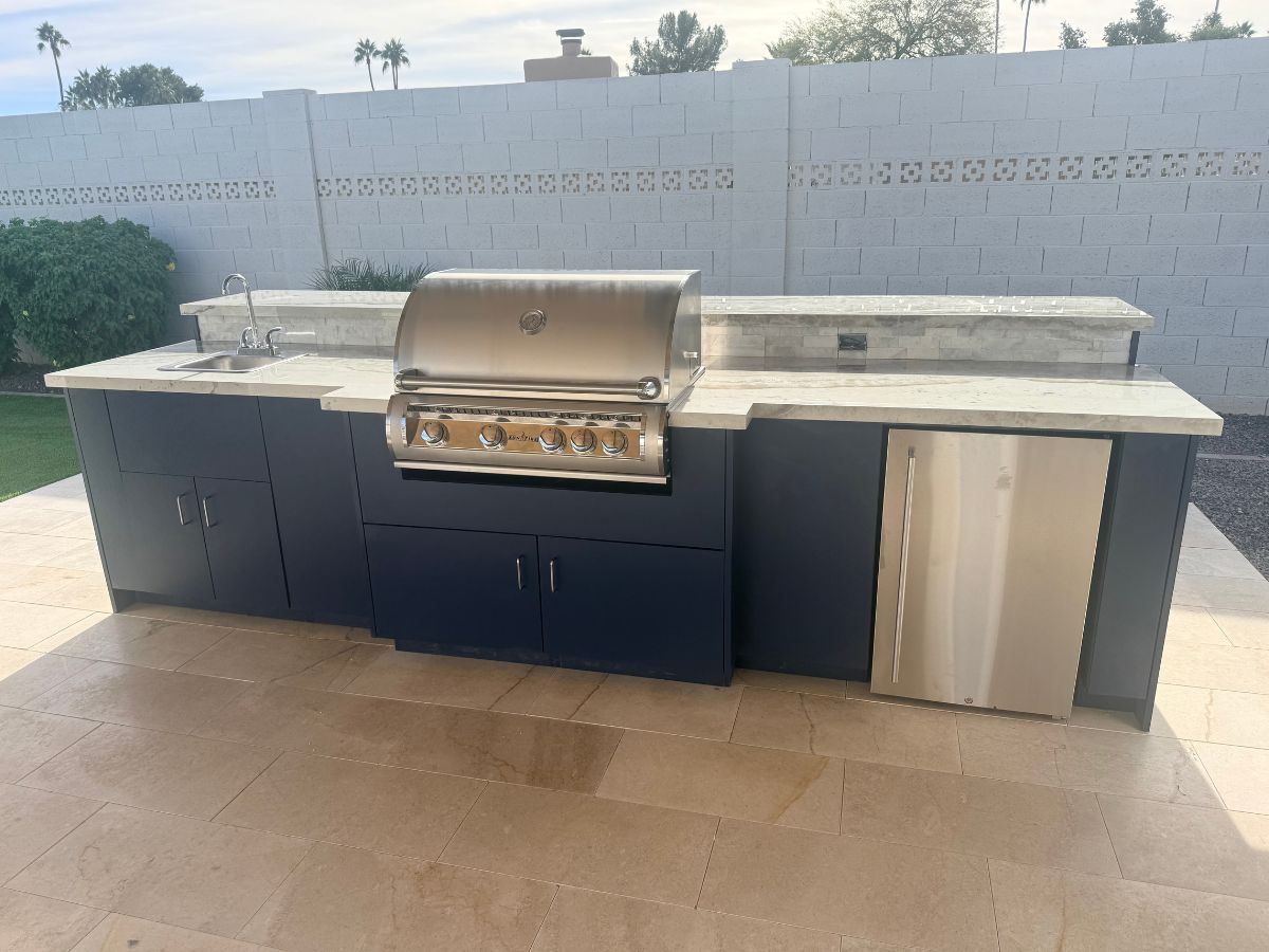 Modern outdoor kitchen island with built-in stainless grill, sink, stone countertop, navy cabinets and fridge on patio