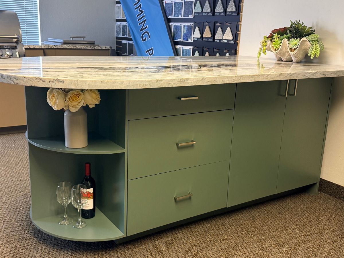Green-cabinet kitchen island with marble countertop, wine bottle and glasses on curved shelves, decorative vase of roses