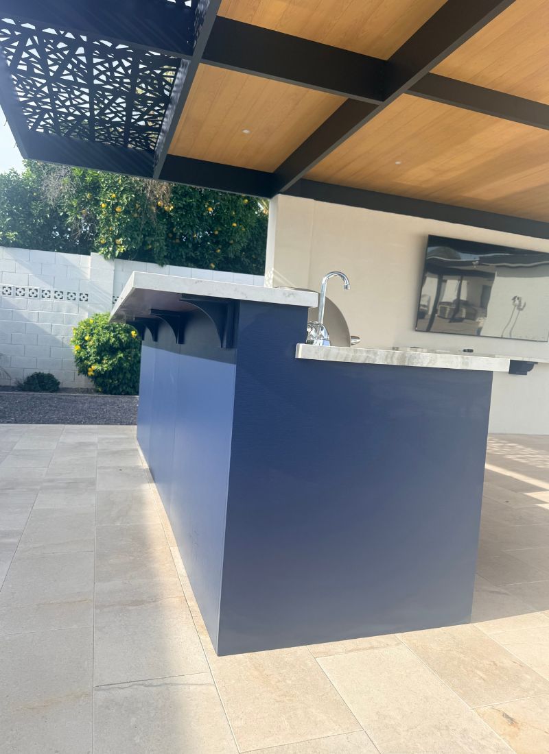 Modern outdoor kitchen island: navy blue base, marble countertop, faucet under wood pergola on tiled patio