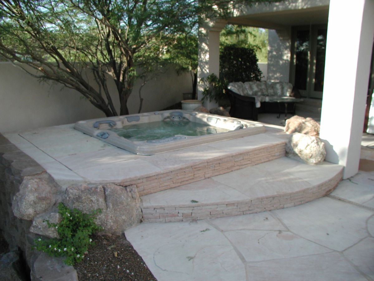 Raised stone patio with sunken hot tub spa, curved steps, desert landscaping and covered seating area in backyard.
