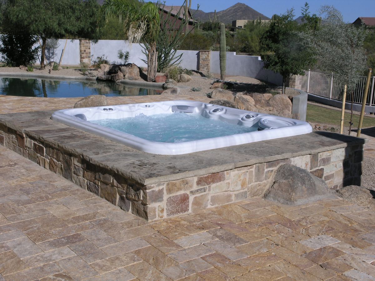In-ground hot tub on raised stone patio beside pool in desert backyard with palm and cactus