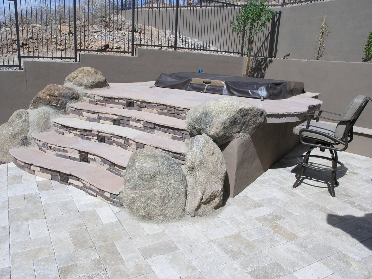 Backyard hot tub with cover on stone-veneered steps flanked by boulders, tiled patio and bar stool