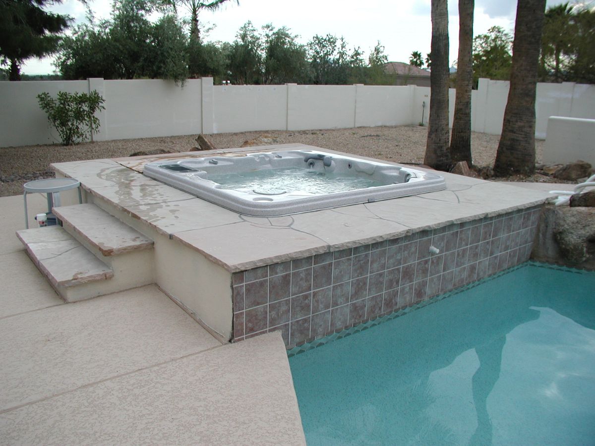 Raised tiled hot tub spa overlooking turquoise swimming pool in a desert backyard with palm trees.
