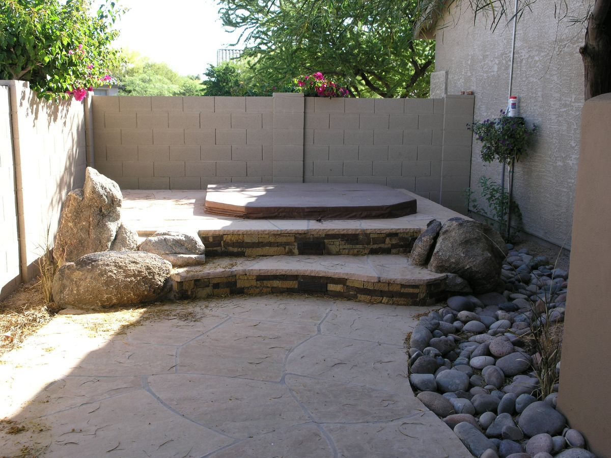 Backyard patio with covered hot tub on raised stone deck, curved flagstone steps, boulders and river-rock landscaping