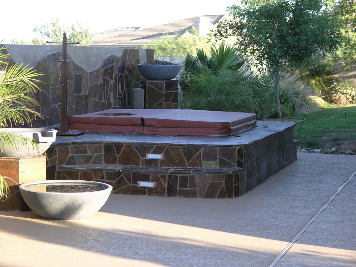 Backyard covered hot tub on stone raised platform with steps, sunlit patio and potted plants