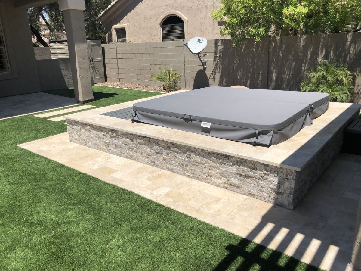 Raised hot tub with gray cover on stone-clad platform, travertine patio and artificial turf backyard