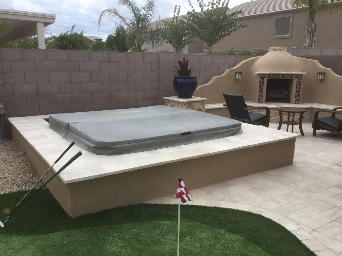 Backyard stone patio with built-in covered hot tub, outdoor fireplace, seating and small putting green with flag.