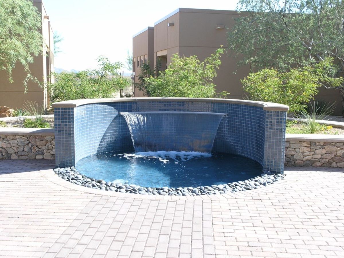 Blue-tiled semicircular waterfall fountain in courtyard with pebble edging, paved plaza and desert landscaping