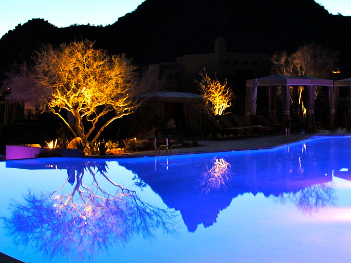 Night resort pool with blue-lit water, illuminated desert trees and mountain silhouette reflection
