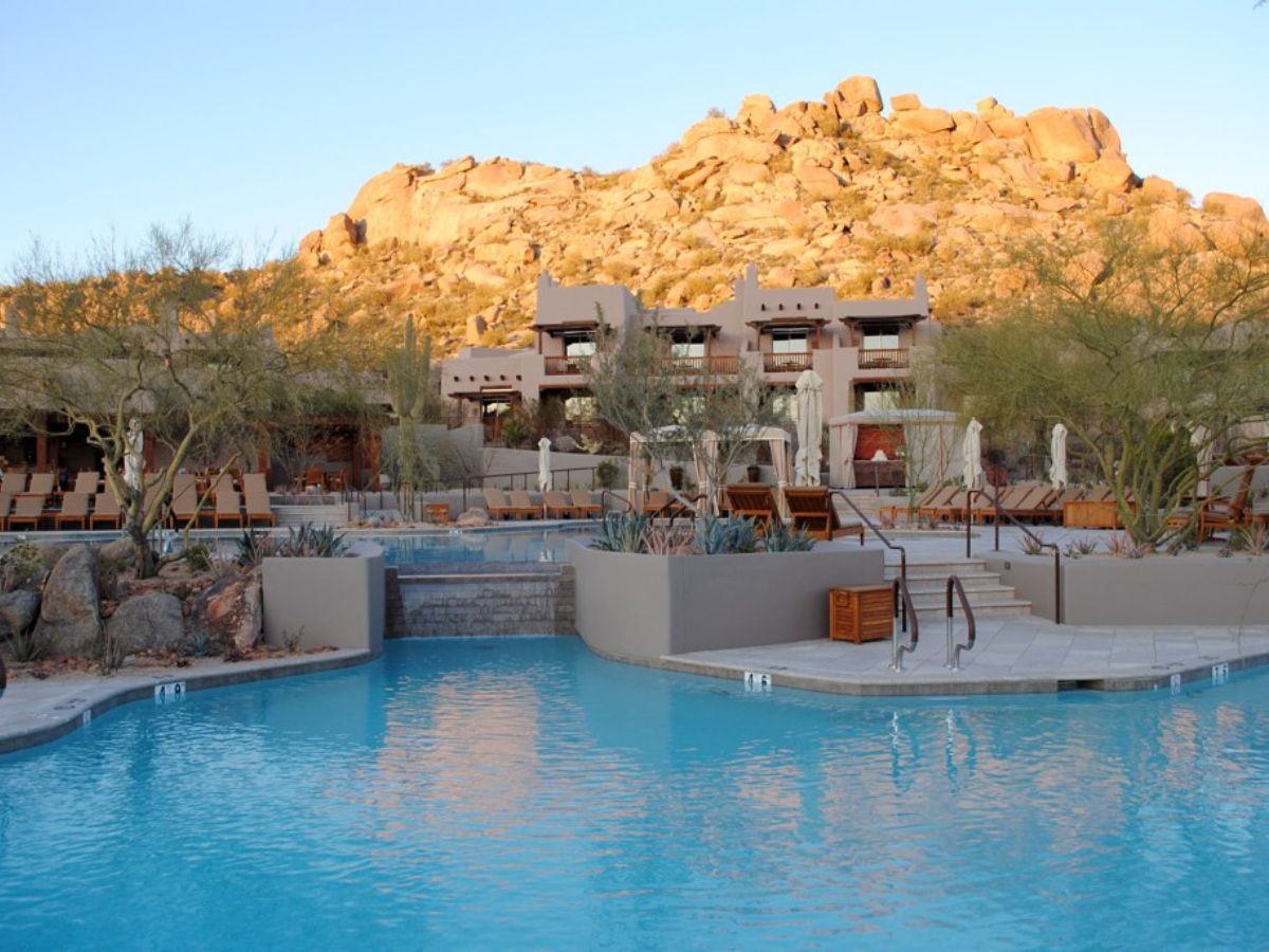Luxury desert resort pool with sunlit rocky hillside, adobe-style suites and lounge chairs at sunset.
