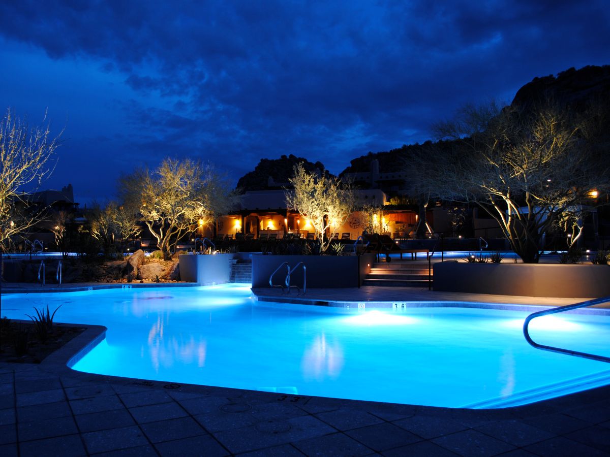 Blue-lit luxury resort swimming pool at night with illuminated trees and a lit lounge area