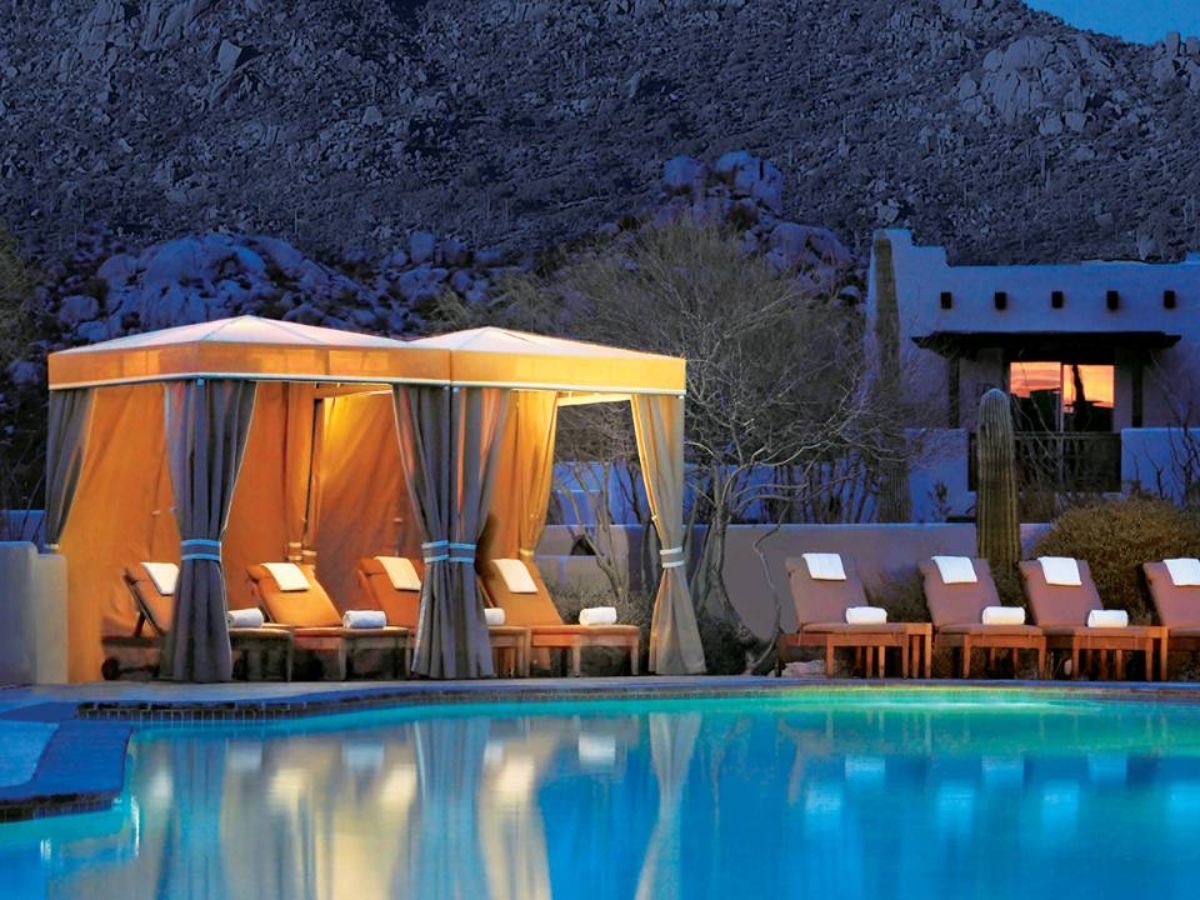 Luxury resort swimming pool at dusk with lit cabana and row of chaise lounges set against desert mountain backdrop