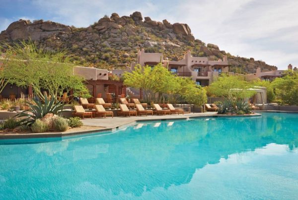 Luxury resort pool with blue water, wooden lounge chairs, desert plants and rocky mountain backdrop
