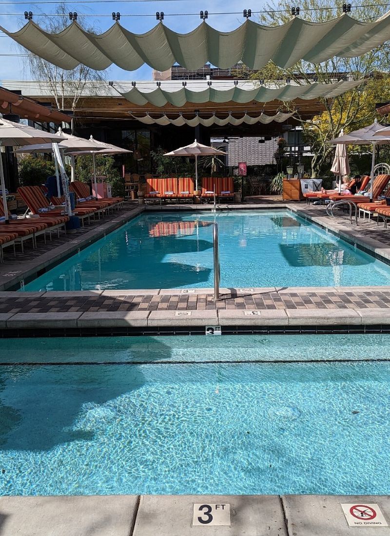 Outdoor resort pool with striped lounge chairs, umbrellas and overhead shade sails, clear blue water and 3 ft shallow end