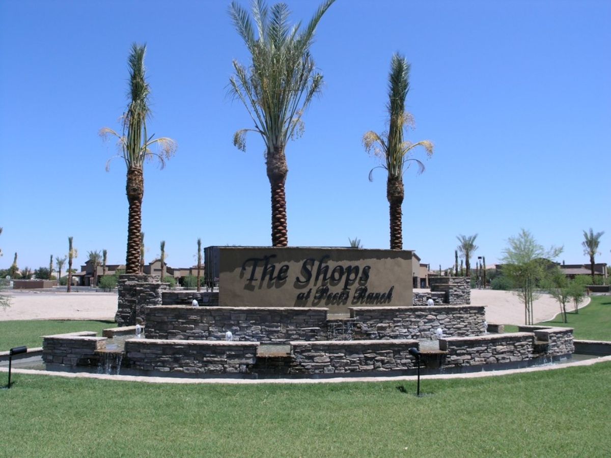 Stone fountain entrance sign reading 'The Shops' with palm trees and manicured lawn under clear blue sky