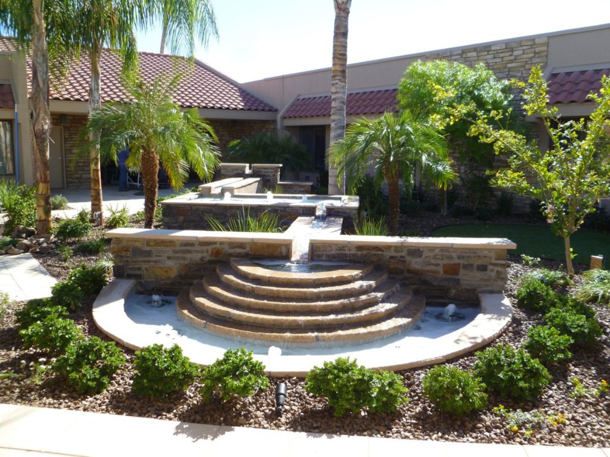 Tiered semicircular stone water fountain in landscaped courtyard with palm trees and Spanish tile-roofed building