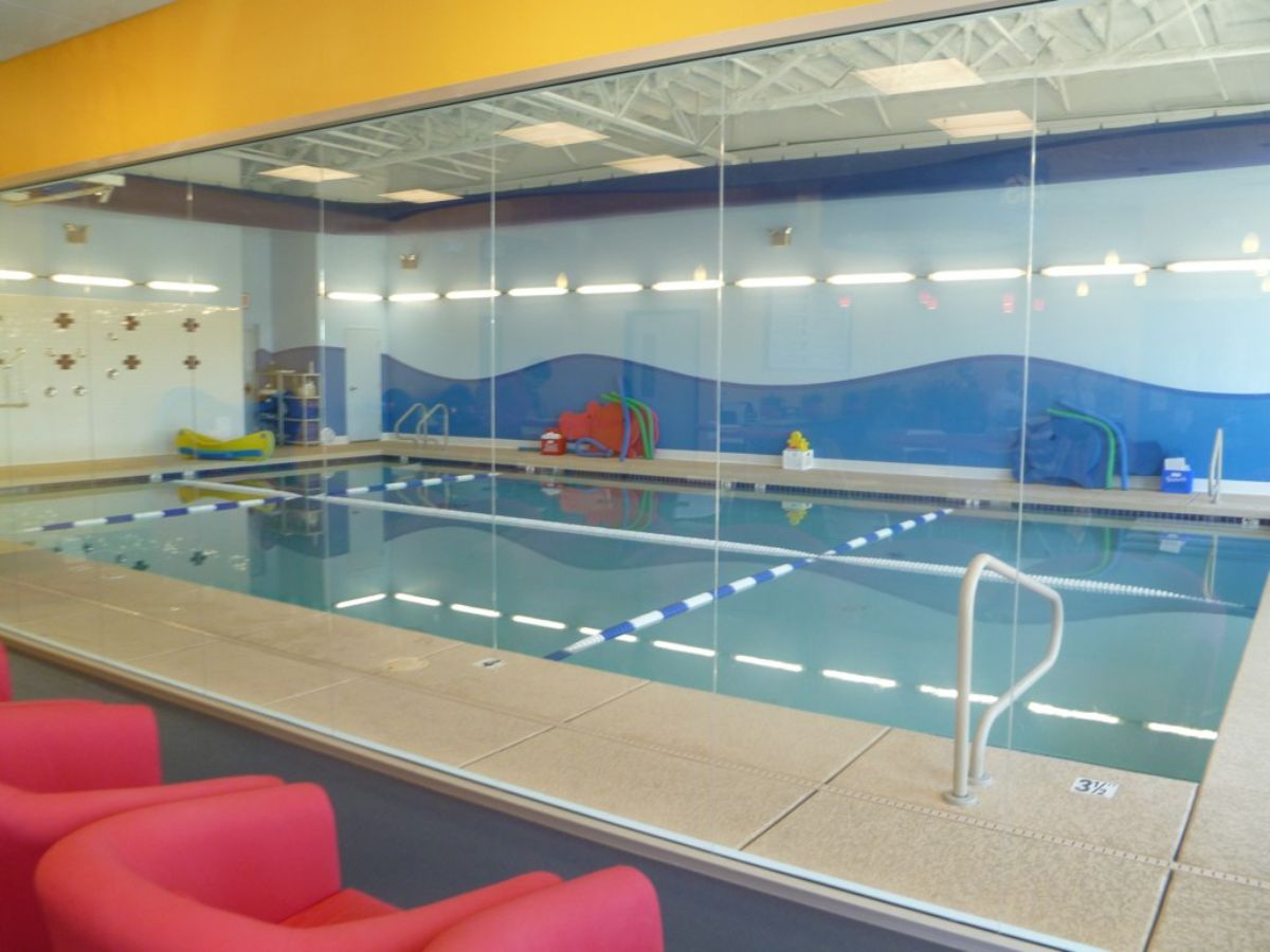 Indoor swimming pool with lane ropes and children's play equipment, viewed through glass with red seats.