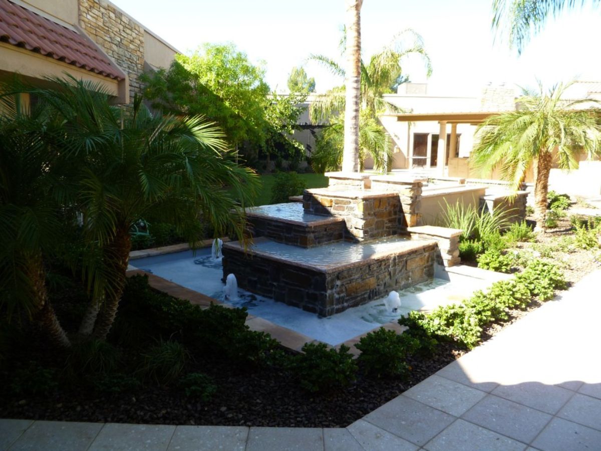 Tiered stone water fountain in a landscaped Mediterranean-style courtyard with palm trees and tiled patio