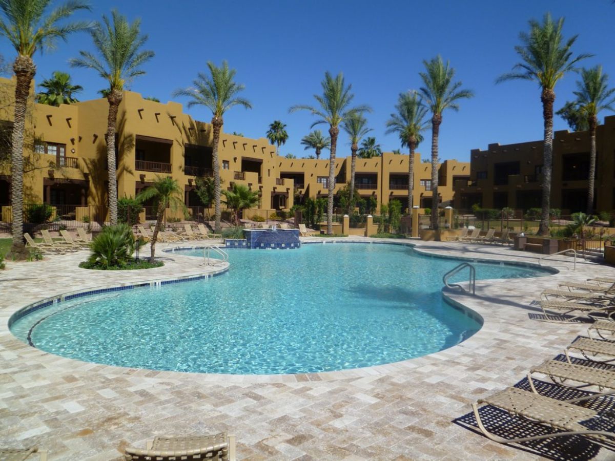 Resort pool with palm trees, lounge chairs and adobe-style hotel condos, sunny vacation oasis under clear blue sky