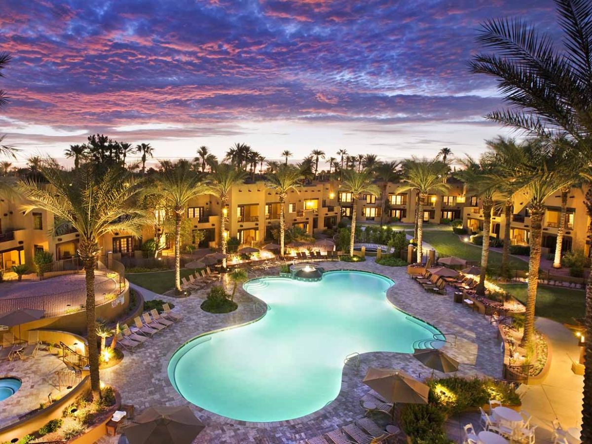 Luxury resort pool at sunset with palm trees, lit hotel rooms and lounge chairs, vacation oasis