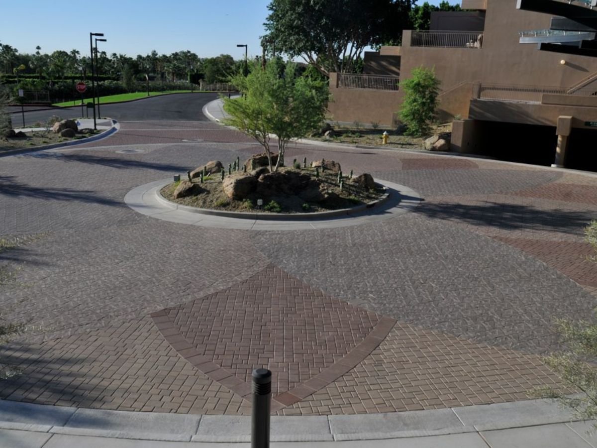 Roundabout with landscaped rock island, trees and cacti, brick-paver roadway to residential building and parking entrance.