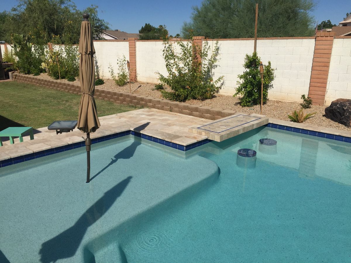 Backyard swimming pool with built-in submerged bar stools, closed umbrella on tanning ledge and travertine patio