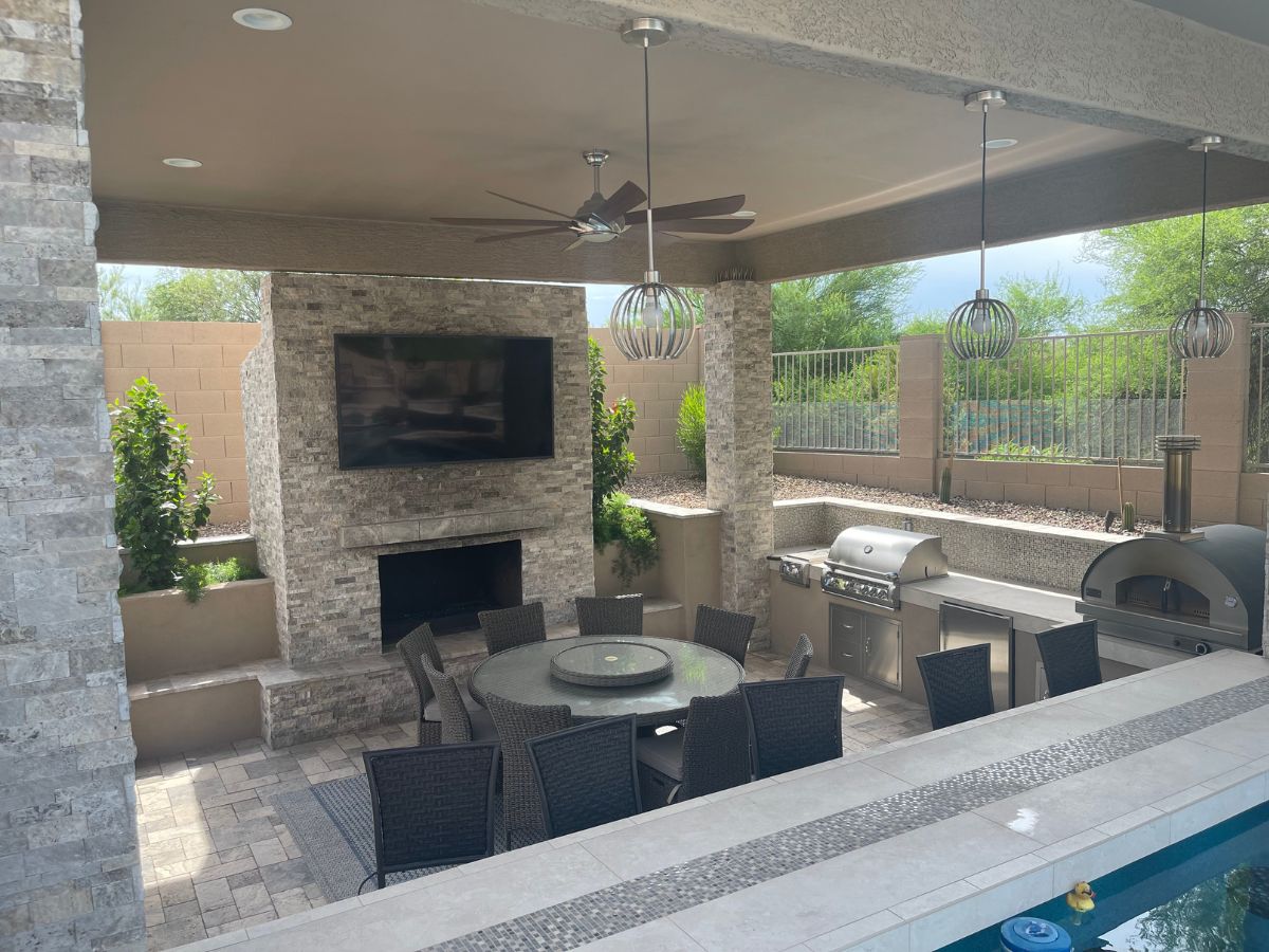 Modern covered outdoor patio with stone fireplace and TV, round dining table, built-in stainless grill and pizza oven by pool.