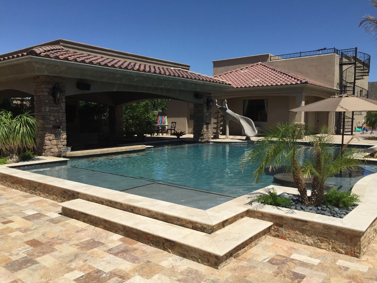 Luxury backyard pool with slide, covered patio, stone tile deck, palm plants and Mediterranean-style home
