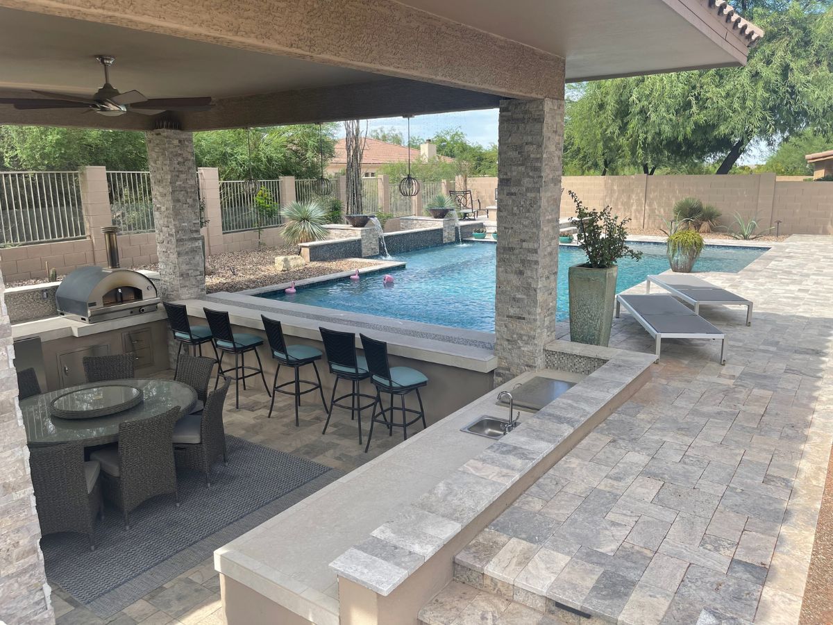 Luxury backyard pool with covered outdoor kitchen and bar seating, stone paver patio, loungers and water features