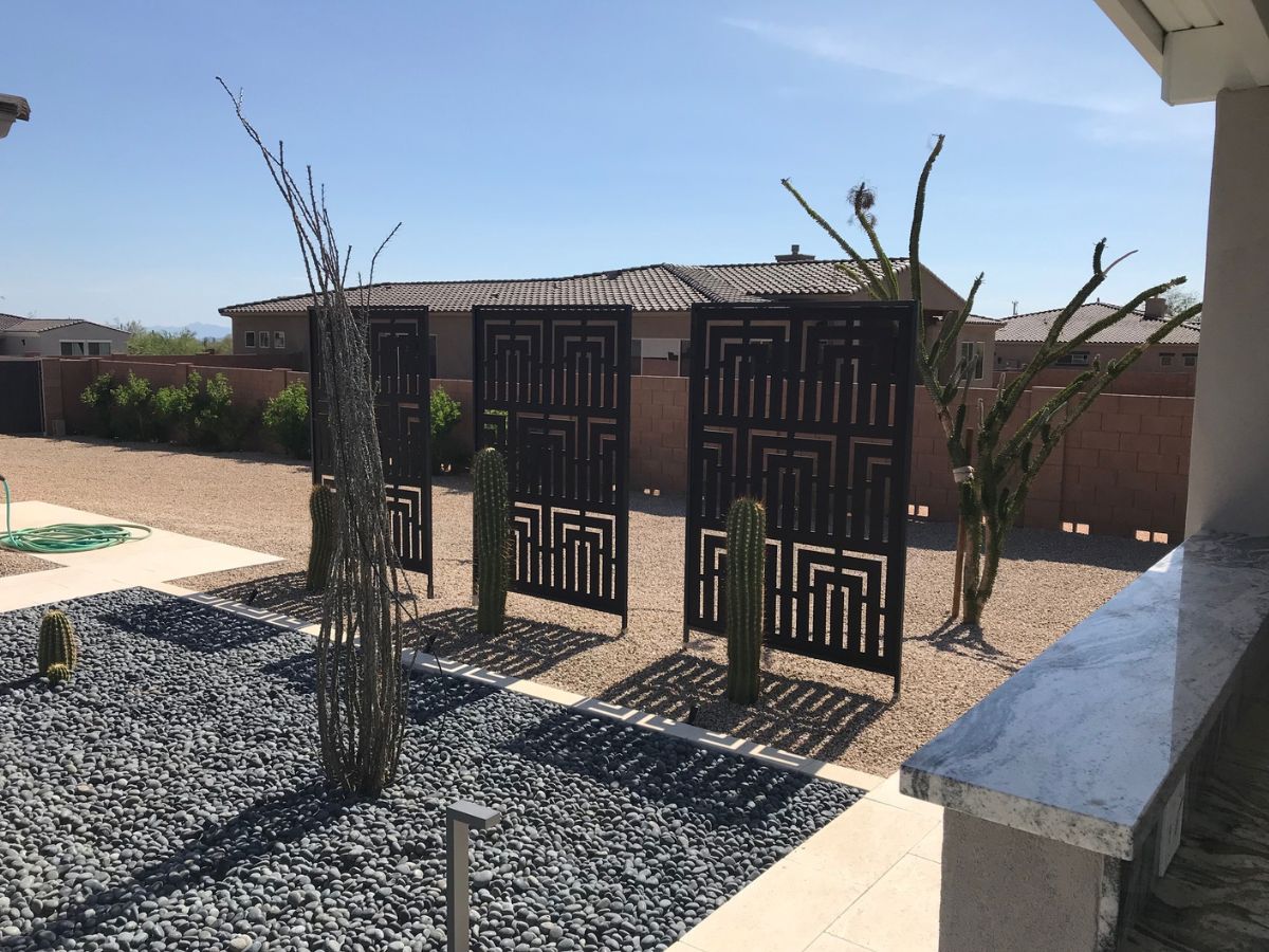 Modern desert backyard with metal privacy panels, tall cacti, and black river-rock xeriscape under clear blue sky