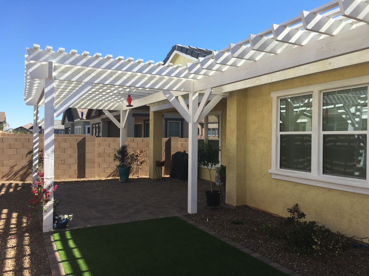 White pergola patio cover over paver patio in sunny backyard beside yellow stucco house with artificial lawn