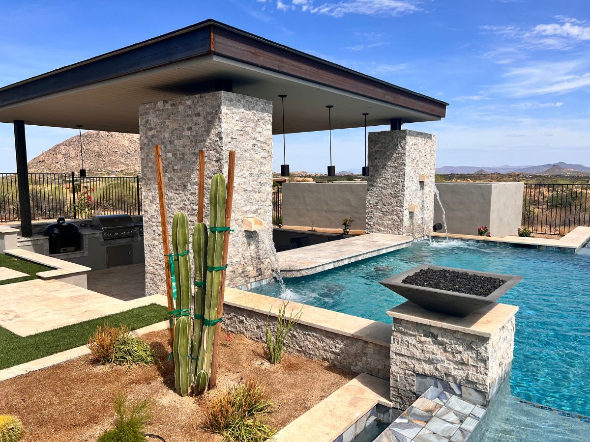Luxury desert backyard with modern pool, stone water-feature pillars, outdoor kitchen under pergola and tall barrel cactus.
