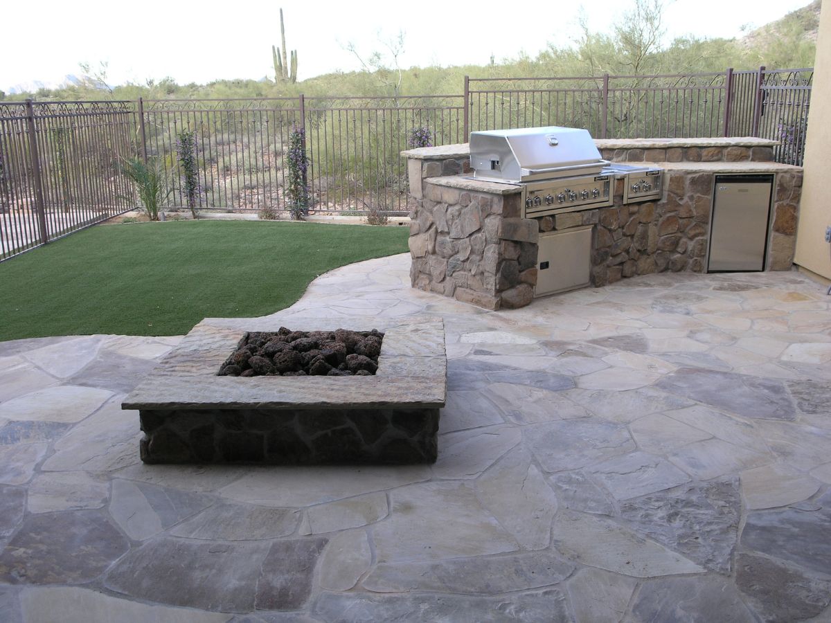 Stone patio with built-in stainless steel grill, outdoor kitchen island and square stone fire pit beside artificial turf.