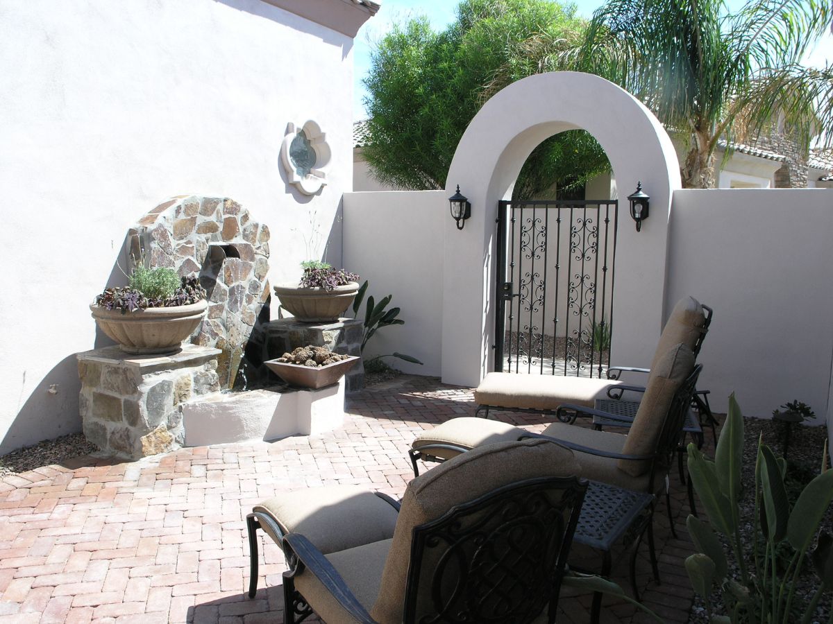 Sunlit Mediterranean patio with stone fountain, potted plants, cushioned lounge chairs and wrought-iron arched gate