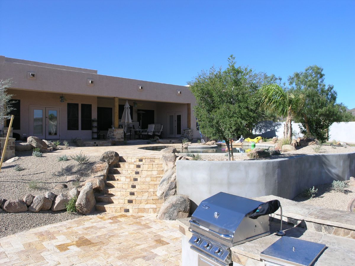 Sunlit desert backyard with stone patio, built-in stainless grill, steps to pool and covered patio seating