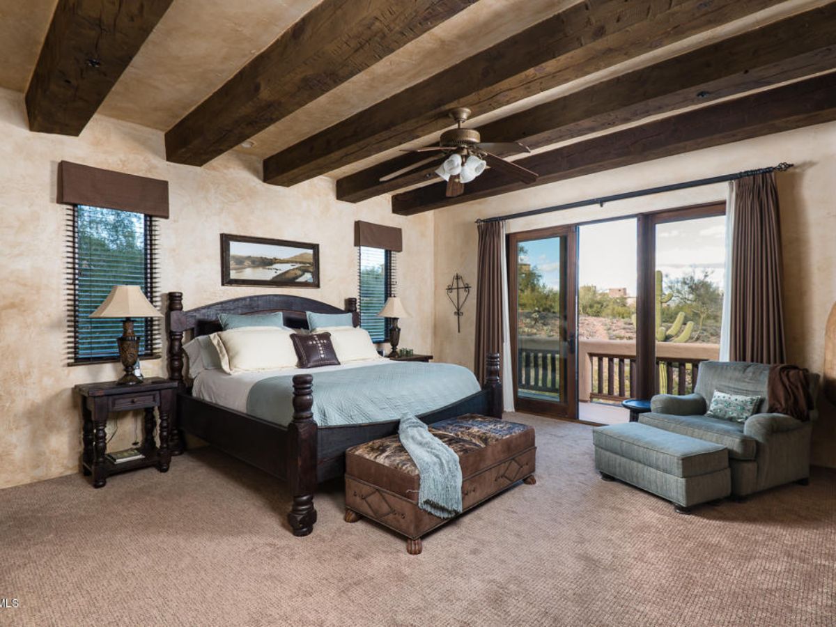 Rustic master bedroom with exposed wood beams, king bed, ottoman, lounge chair and sliding glass doors to desert view.