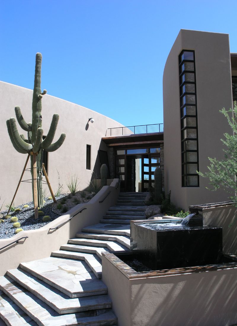 Modern desert home entrance with saguaro cactus, tiered stone steps, stucco walls and black water fountain under blue sky
