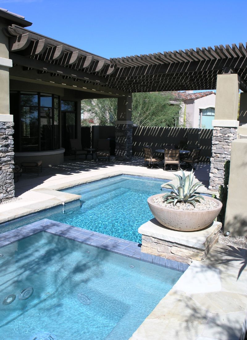 Luxury backyard pool and spa with pergola-covered patio, stone columns and agave planter under clear blue sky