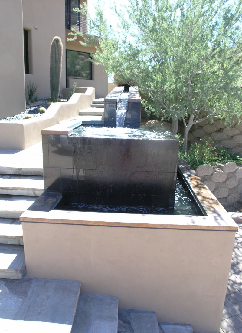 Modern tiered black-stone water feature with reflecting pools beside stucco steps, desert landscaping with cactus and trees