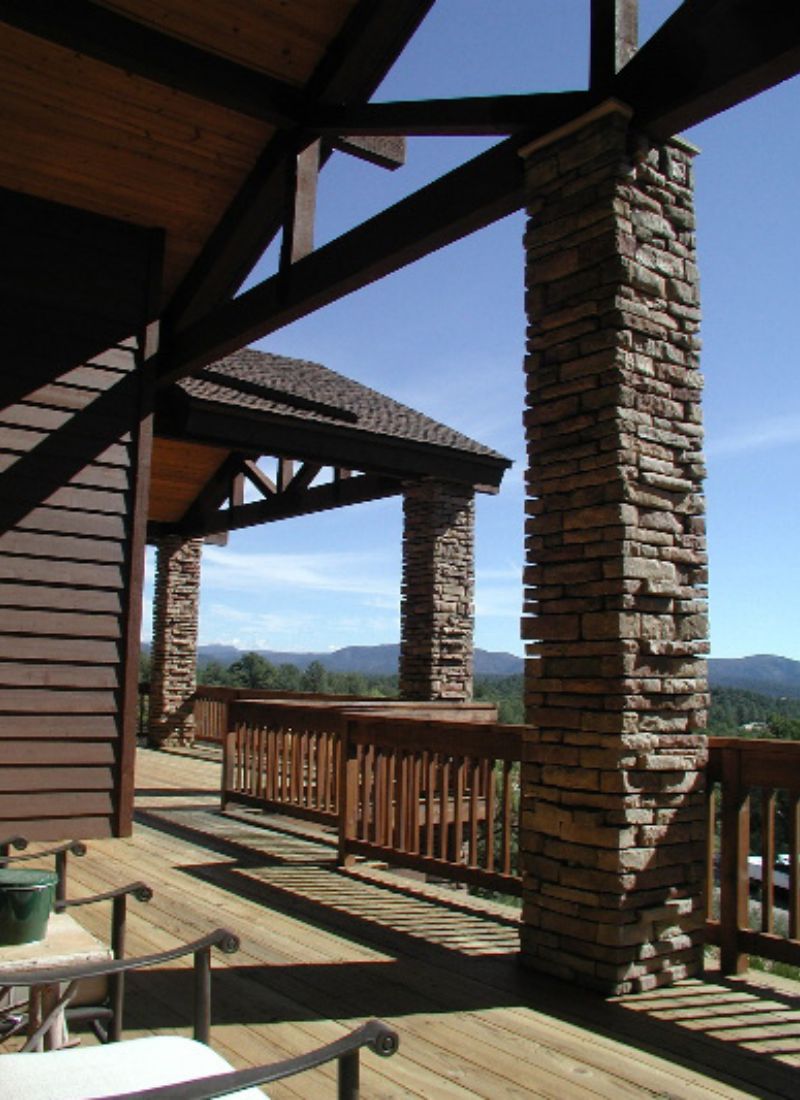 Mountain cabin porch with stacked stone pillars and wooden deck railing, scenic forested hills and blue sky view