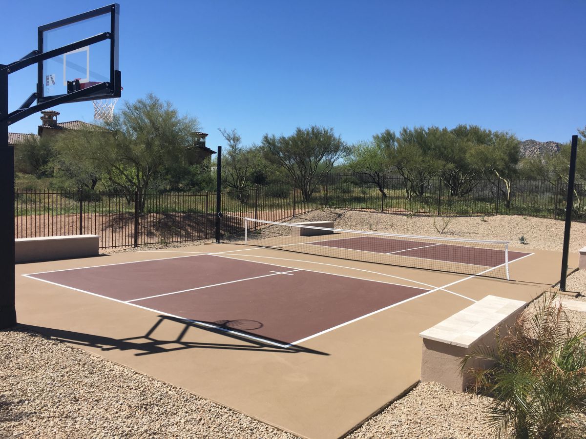 Residential backyard pickleball court with basketball hoop, desert landscaping and iron fence under clear blue sky