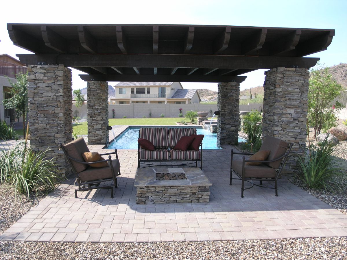 Backyard pergola with stacked stone pillars, pool view, striped outdoor sofa and chairs around stone fire pit on paved patio
