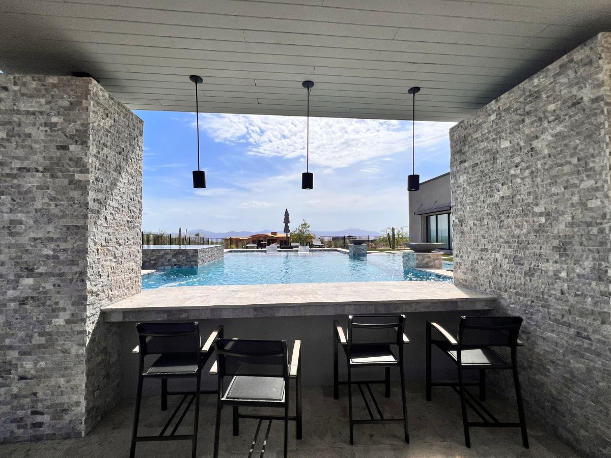 Luxury poolside bar with four stools, hanging pendant lights, stone walls and infinity pool overlooking desert mountains