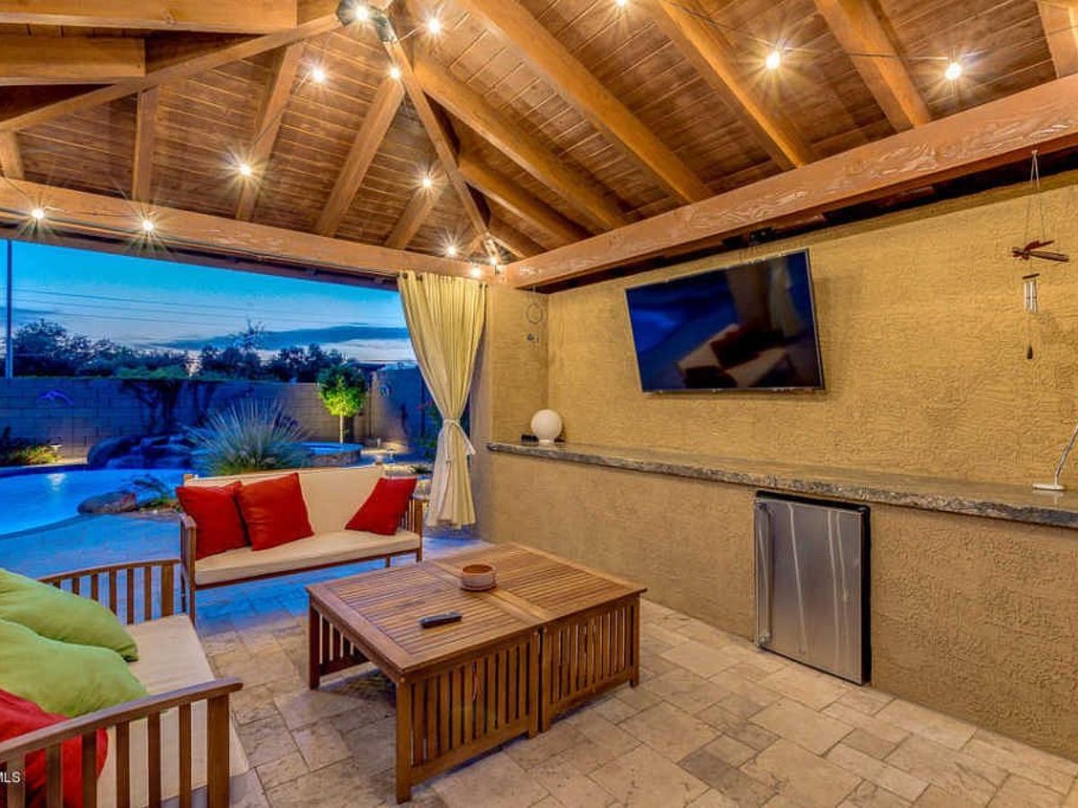 Covered patio with wooden vaulted ceiling, outdoor seating, wall-mounted TV and pool view at dusk