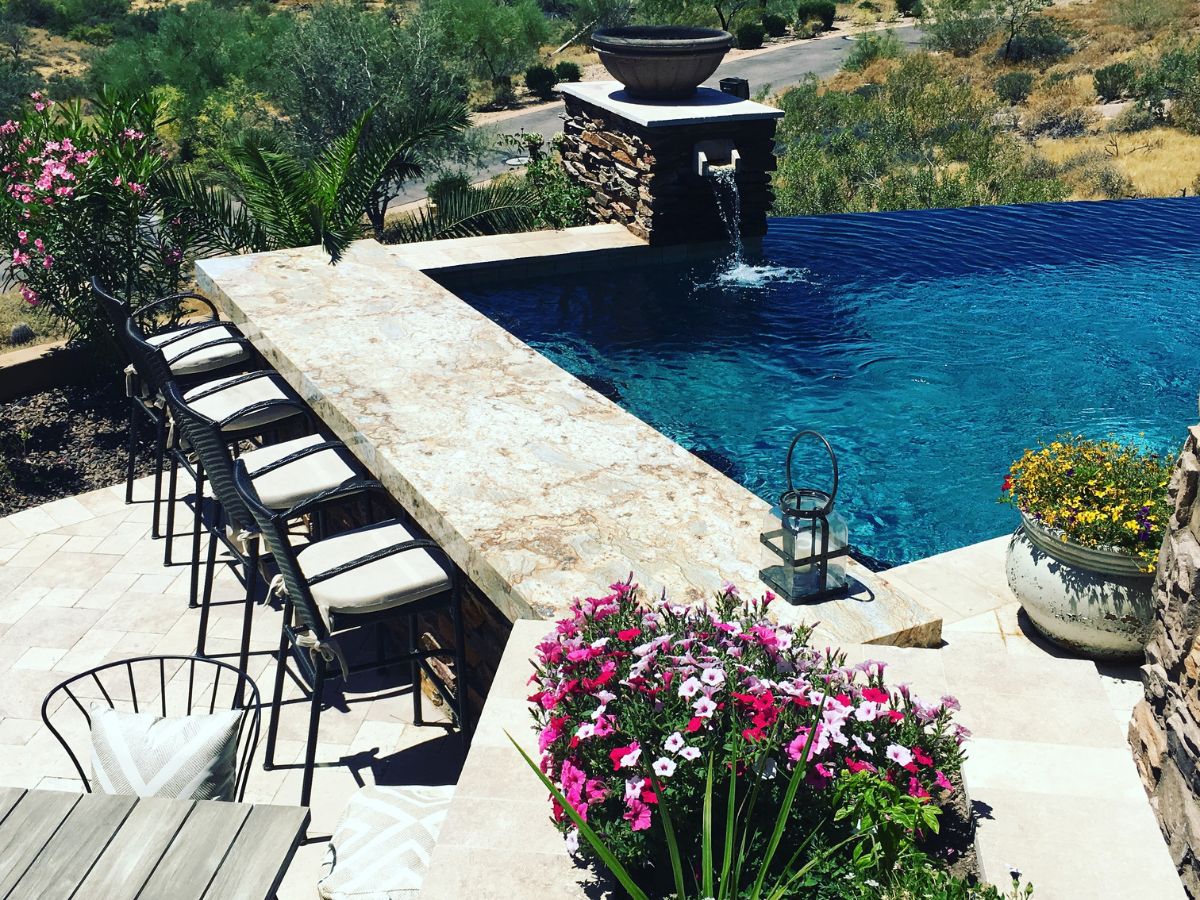 Luxury backyard infinity pool with stone bar and cushioned bar stools, vibrant pink flowers and desert landscaping.