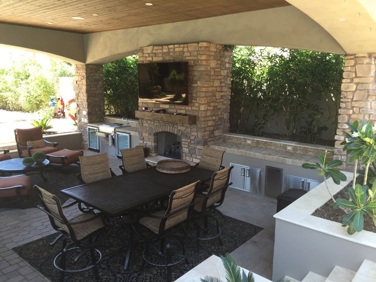 Covered luxury outdoor patio with stone fireplace, wall-mounted TV, large dining table, wicker chairs and outdoor kitchen.