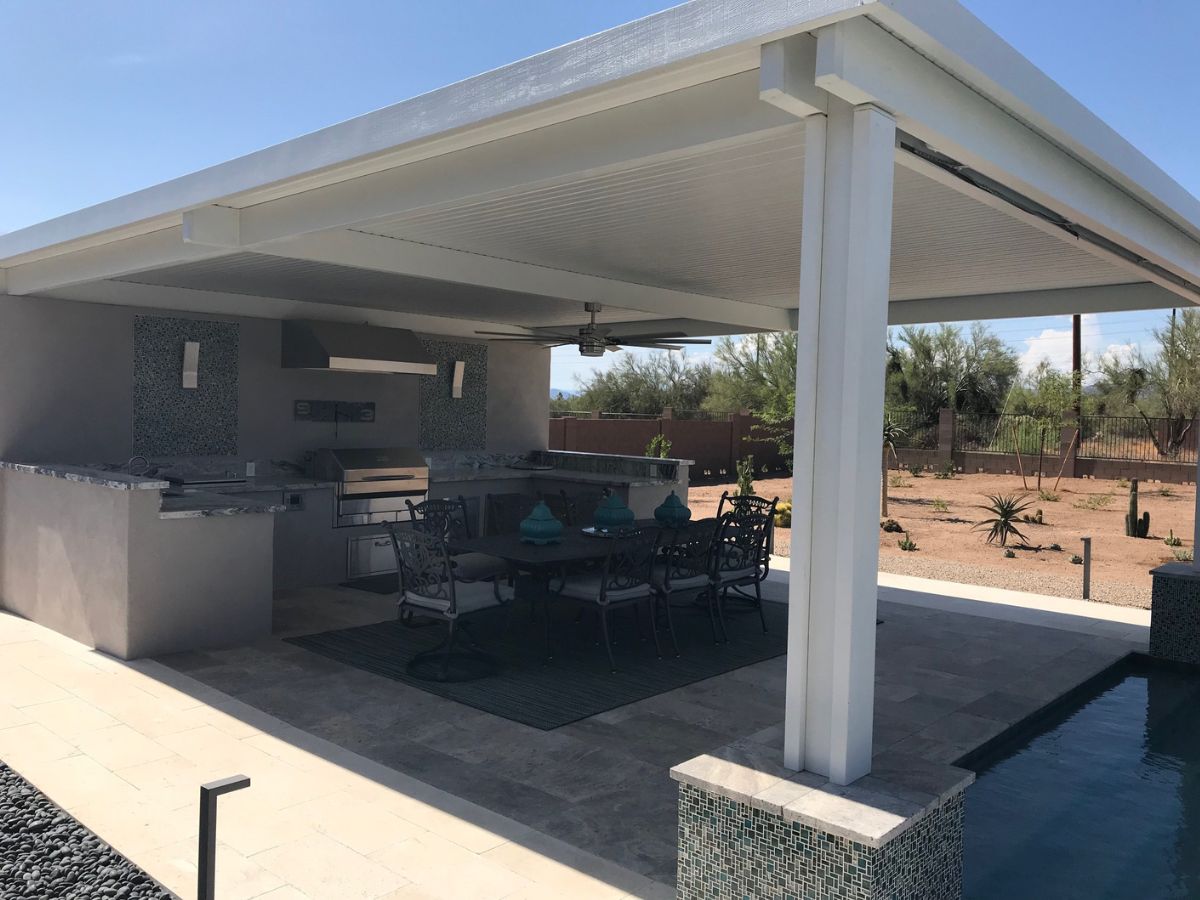 Covered outdoor kitchen patio with built-in stainless grill and dining set beside pool in desert backyard