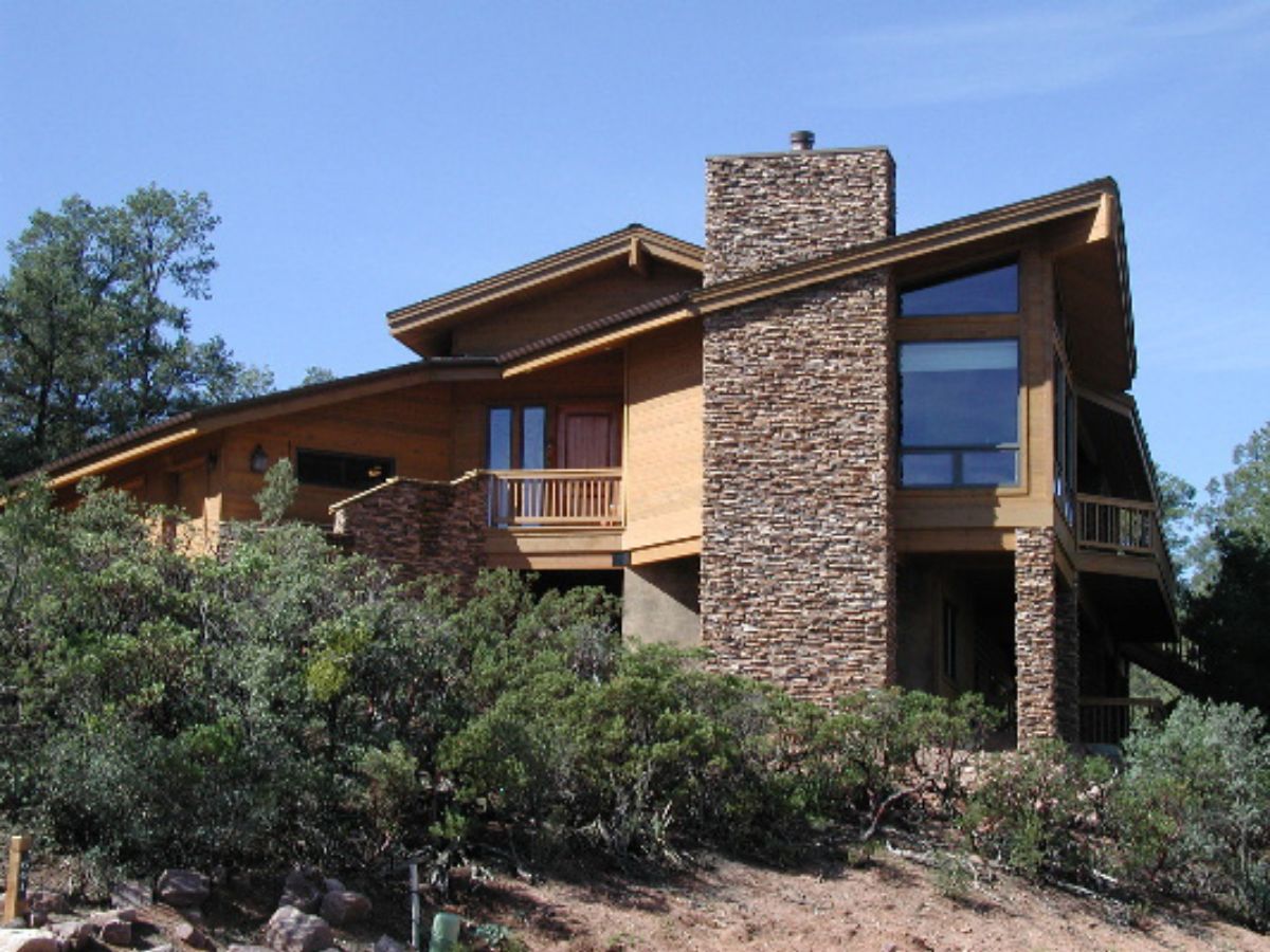 Modern mountain house with wood siding, stone chimney, large windows and balcony above scrubby hillside under blue sky