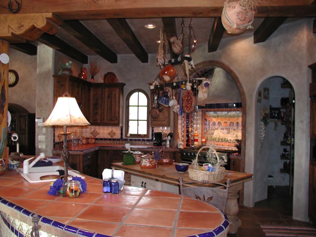Rustic Mediterranean kitchen with terracotta tile countertops, wooden ceiling beams, hanging pots and tiled backsplash.